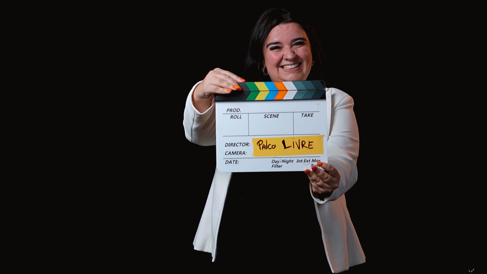 Heloísa de blazer branco segurando a claquete Palco Livre, à direita sobre fundo preto